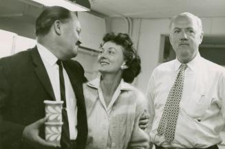 A photograph of three people - two men and a woman between them - standing in a room. They are …