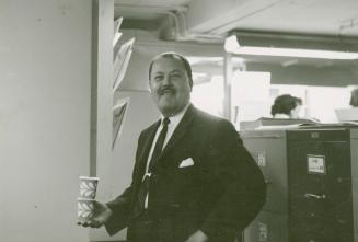 A photograph of a man wearing a business suit and tie standing in an office in front of some fi…