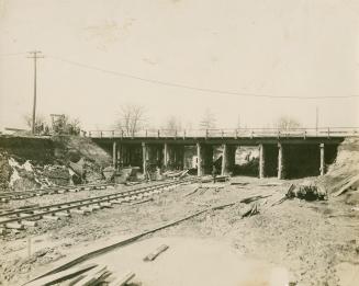 A photograph of a subway line under construction, passing under a railway bridge. There are wor…