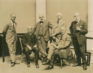 A photograph of six men sitting and standing in a room with framed pictures propped against the…