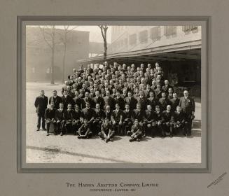 A group staff photograph comprised of several dozen men sitting and standing in front of a buil…