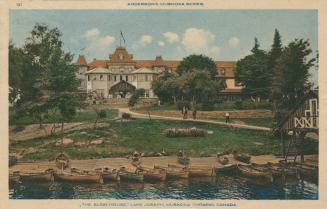 A large resort hotel in the background. Canoes docked at a shoreline in the foreground.