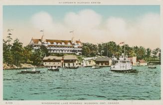 A steam ship and canoes on a lake in front of huge hotel complex and out buildings.