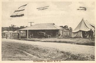 Sepia toned picture of two planes flying over buildings and tents in an airfield.