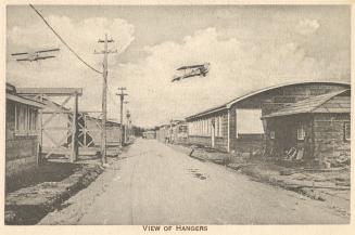 Sepia toned picture of two planes flying over hangar buildings.