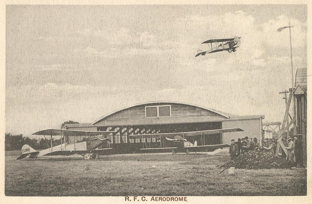 Sepia tone picture of two airplanes on the ground in front of a large, commercial building. Ano…