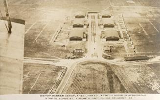 Black and white shot from a flying plane of military planes lined up on each side of six hanger…