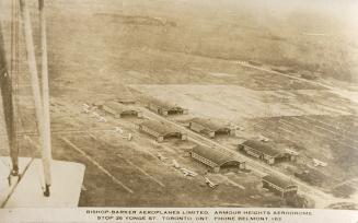 Black and white shot from a flying plane of military planes lined up on each side of six hanger…