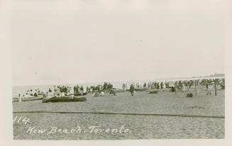 Black and white picture of a crowd of people on a sandy beach to the right of a large body of w…