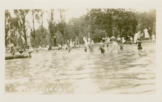 Black and white picture of a crowd of people splashing the water beside a sandy beach.