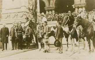 Black and white photograph of men standing and on horseback in front of Romanesque arches.