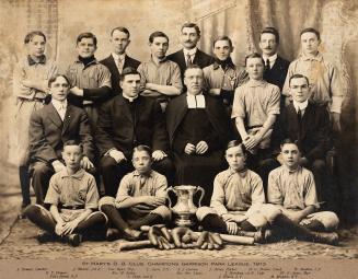 A photograph of a baseball team posing with bats, gloves, a ball and a trophy in a photo studio…