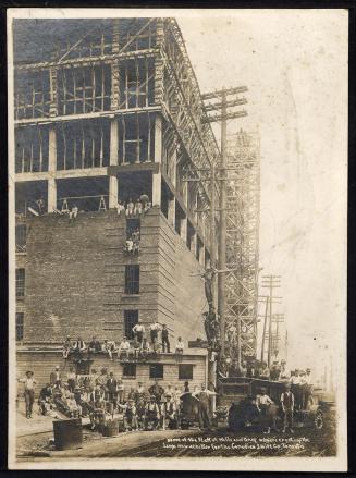 A photograph of a large group of construction workers posing in front of a partially-built eigh…