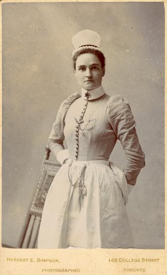 A posed photograph of a young woman wearing a nurse's uniform. She is standing in front of a ro…