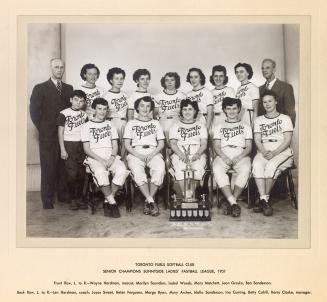 A photograph of a women's softball team posing in front of a trophy. They are sitting and stand…