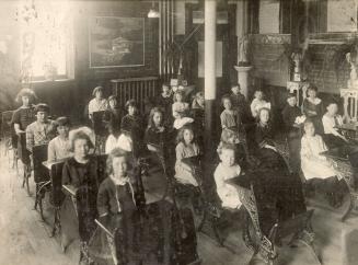 A photograph of approximately twenty young students, boys and girls, sitting at wooden desks in…