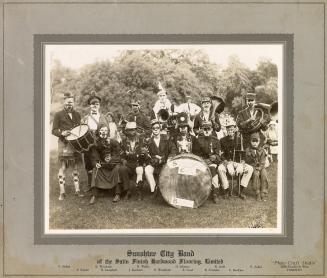 A photograph of the members of a band posing on a grassy lawn in front of some trees and bushes…