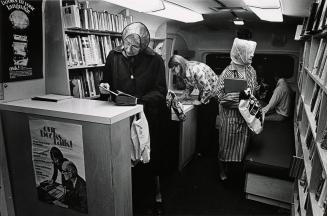 Shows a number of people using a Bookmobile. An older woman in a rain hat looks at a hardcover …