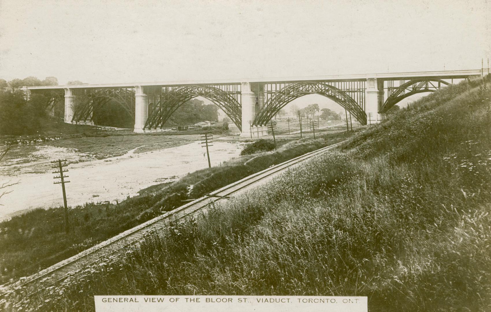 Black and white photograph of a large bridge spanning a valley with railroad tracks visible.