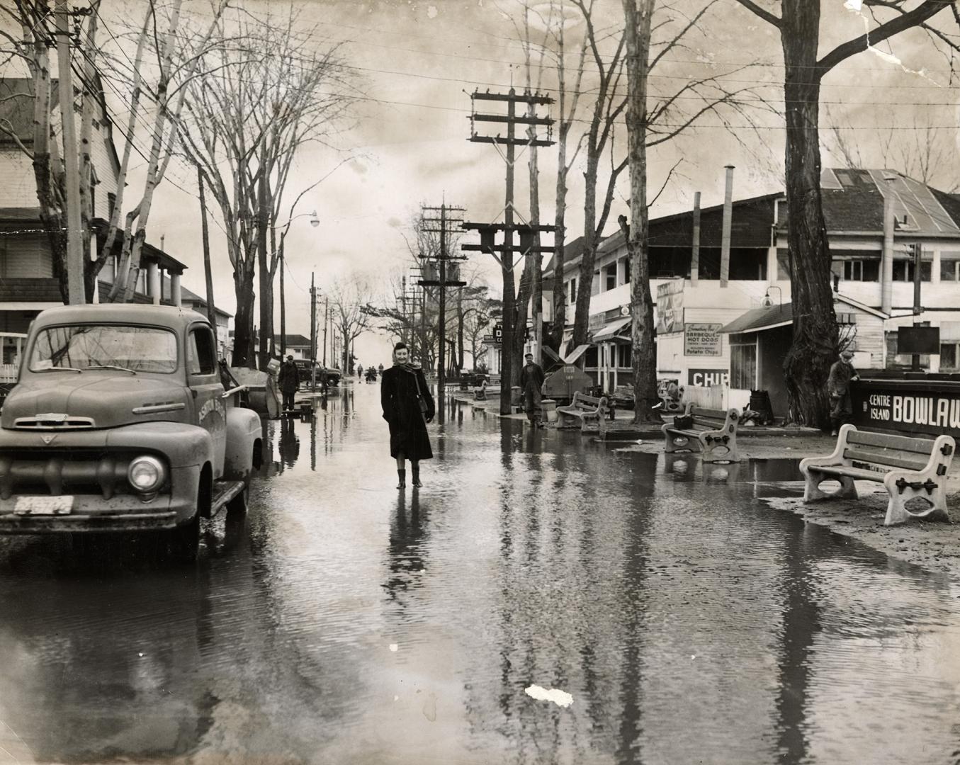 Picture of a woman walking down flooded street with stores on both sides and a truck parked on …