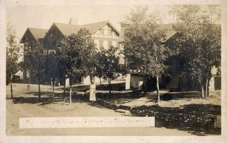 Black and white photograph of a nurse standing on the lawn in front of a large mock-Tudor house…