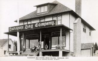 Black and white photograph of three people sitting on a verandah in front of a store. Sign says…