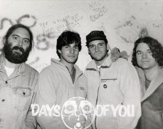 A photograph of a musical group posing in front of a wall with graffiti on it, and the band nam…