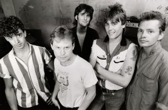 A photograph of a musical group posing in front of musical equipment including a speaker and tu…