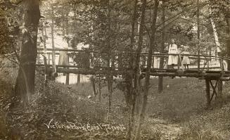 Black and white photograph of three women standing on a log bridge in a wooded area.