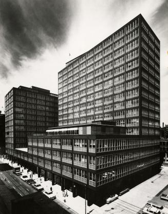 A photograph of a large split-level office building, located at the corner of a city street. Th…