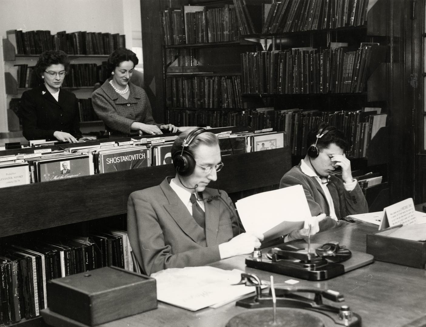 Photo of two people in a library listening to records at a table and two others standing lookin…