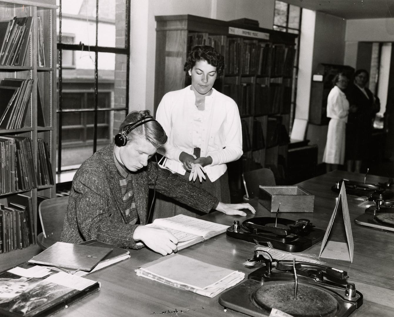 Photo of man at a table with turntable listening to a record and a woman watching. 