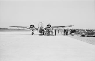 A photograph of a twin-propeller airplane on the runway of an airport. There are a number of ca…