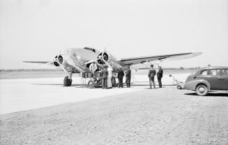 A photograph of a twin-propeller airplane on the runway at an airport. There is a car parked to…