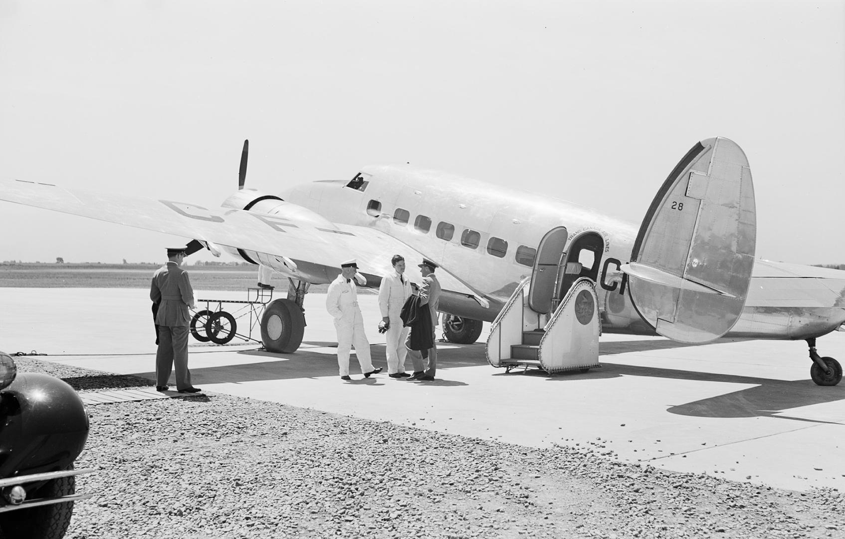 A photograph of a twin-propeller airplane on the runway of an airport. There is a car parked be…