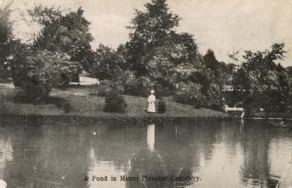 Black and white postcard of a pond surrounded by trees. The caption on the bottom front states,…