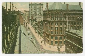 Colorized photograph of an early skyscraper a the bottom of a sprawling city street.