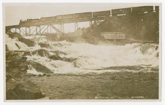 Black and white photograph of a freight train traveling over a bridge which is crossing a water…