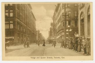 Picture of a street with people lined up on the sidewalk waiting for a street car. 