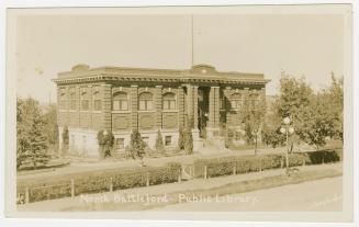 Picture of a two storey library building surround by trees and hedge at front. 