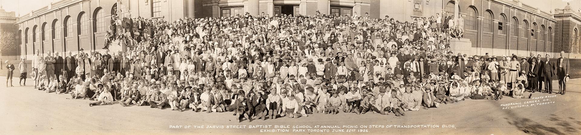 A panoramic photograph of a large group of people posing in front of an office or industrial bu…