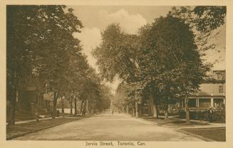 Black and white photograph of a city street bordered by large houses and trees. Coat of arms in…