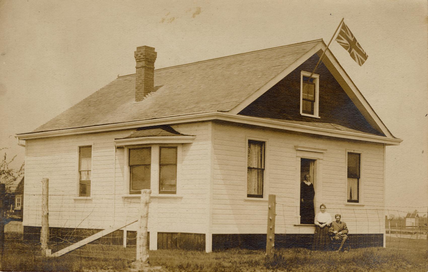 Sepia-toned postcard depicting a home with no entrance steps to the front door, a British flag …