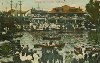 Colorized photograph of a huge crowd of people watching people in canoes in front of a large bu…