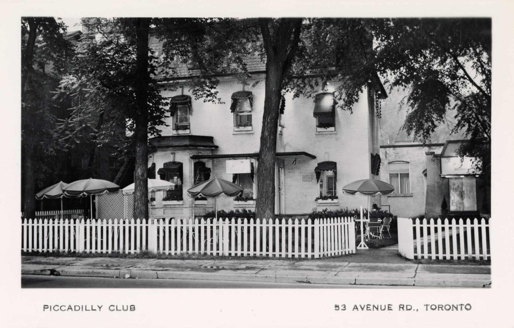 Black and white photo postcard depicting an illustration of a large house with a white picket f…