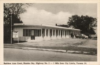 Black and white photo postcard depicting an image of motel with a car and people standing in th…