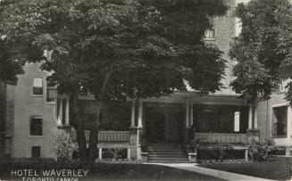 Black and white photo postcard depicting an image of a large house with a front porch being use…