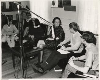 Photo of four young people and two adults sitting talking and large microphones in front of the…