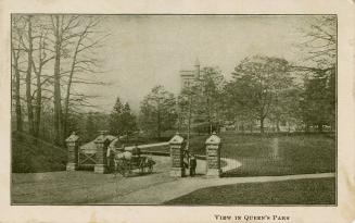 Horse and cart going through brick gates on a roadway in front of trees and a large collegiate …