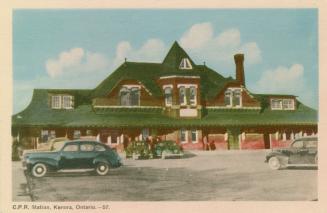 Colorized photograph of cars in front of a one and a half story, Victorian public building.